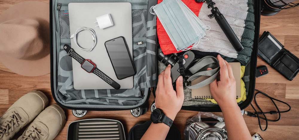 Bild zeigt Hände die einen Reisekoffer packen mit lauter Tech Gadgets wie Smartphone, Kamera, Uhr, Selfie-Stick; Bildquelle: freepik.com/jcomp