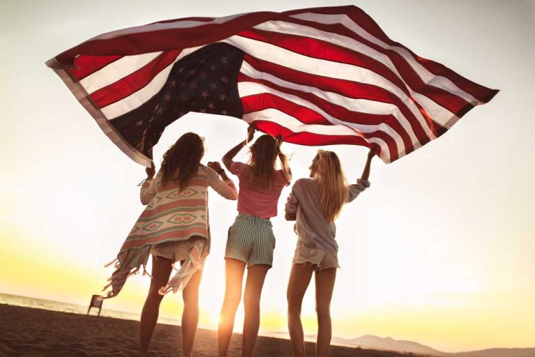 Drei Teenager halten eine amerikanische Flagge am Strand die im Wind weht.